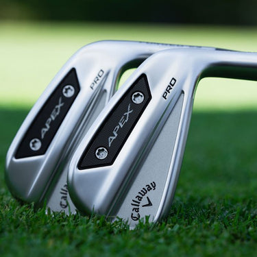 Two Callaway Apex Pro 24 Golf Irons lying toe down on grass with the back of the club heads showing.